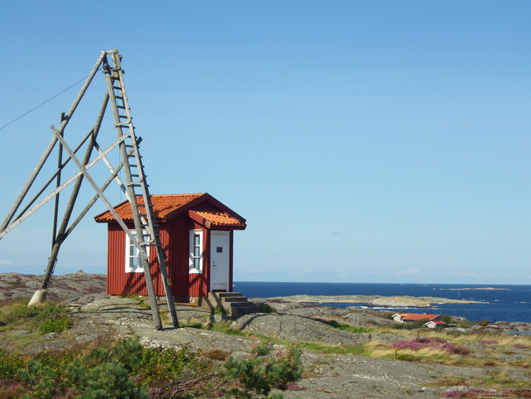 Bild. Litet rött hus med vita knutar högt på berget. Havet i bakgrunden.