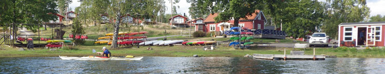 Bild. Kajaker ligger på stranden och på ställning. I ett litet hus står han som hyr ut kajaker. Christer sitter i sin kajak på vattnet.