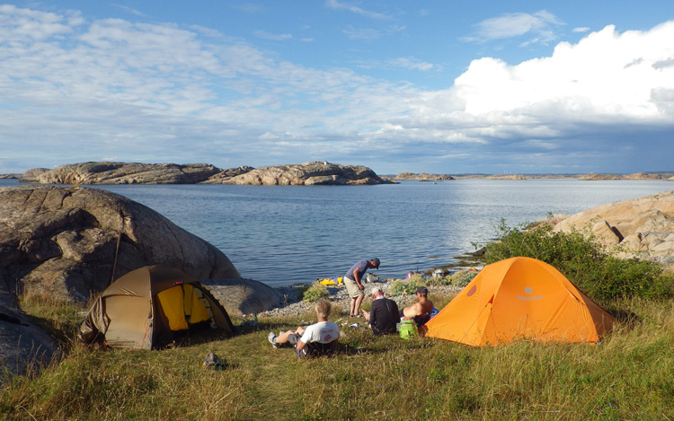 Bild. Tvä tält. Fyra människor. Gräset sluttar mot havet. Klippor på båda sidor. Ö i bakgrunden.