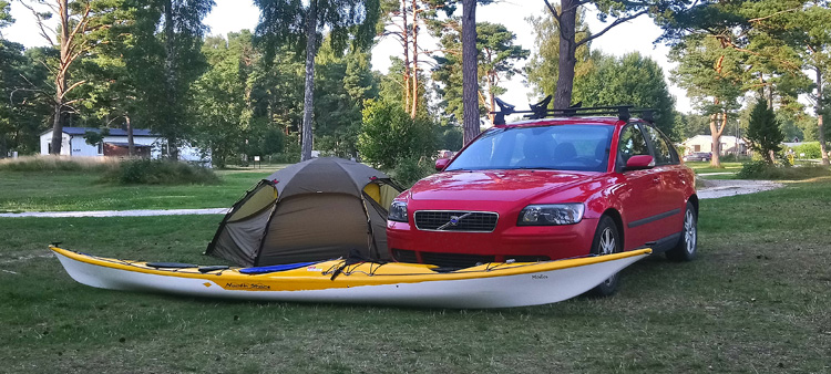 Bild. Kajakaen ligger framför bilen och tältet i gles tallskog.