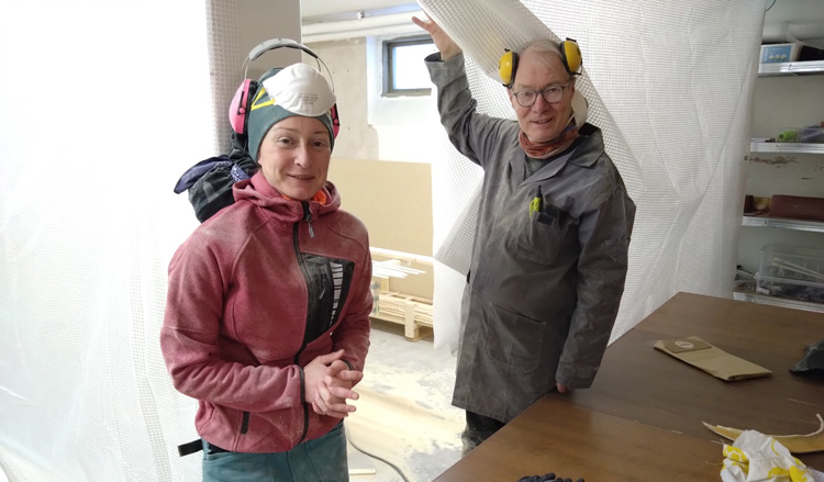 Bild. Linda och Peter i verkstaden fulla av fint sågspånsdamm gläntar på presenningen till byggplatsen.
