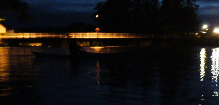 Bild. Mörker. Framför de upplysta broarna vid Skjulsta tecknar sig svagt silhuetten av en kajak.